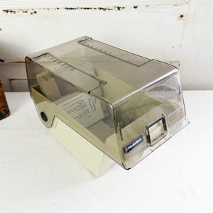 May include: A beige plastic MediaMate storage case with a hinged, translucent dark gray lid. The unit features a rectangular handle on the front and contains a stack of white index cards inside. The design includes a clear top section for viewing.