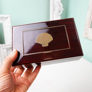May include: A rectangular, wooden jewelry box with a glossy, dark reddish-brown finish. The box features a gold-colored seashell emblem and a gold-colored border. The box is held in a hand.