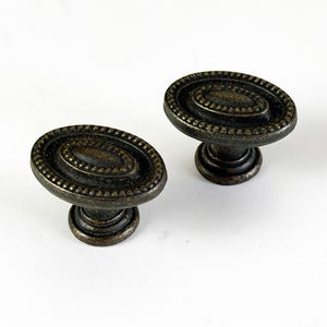 May include: Two antique bronze oval cabinet knobs with a decorative beaded edge and a dark finish. The knobs are on a white background. These knobs are suitable for furniture restoration or adding a vintage touch to cabinets and drawers.
