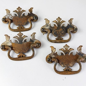 May include: Four matching metal drawer pulls with a weathered, tarnished finish. Each pull features a decorative backplate with an ornate, symmetrical leaf-like design and a curved, swinging handle attached by two small circular mounting points.