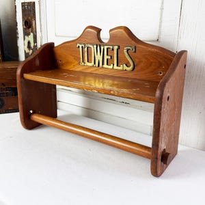 May include: A wall-mounted wooden towel rack featuring a natural brown finish, a flat upper shelf, and a lower horizontal rod for hanging towels. The back panel displays the word TOWELS in raised, metallic, gold-toned lettering centered above the shelf.