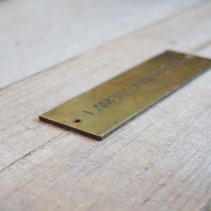 Pode incluir: Uma etiqueta retangular de lat&atilde;o com o texto gravado "LAMBDA DESIGN" e um pequeno furo numa extremidade. A etiqueta est&aacute; colocada sobre uma superf&iacute;cie de madeira clara, criando um contraste.