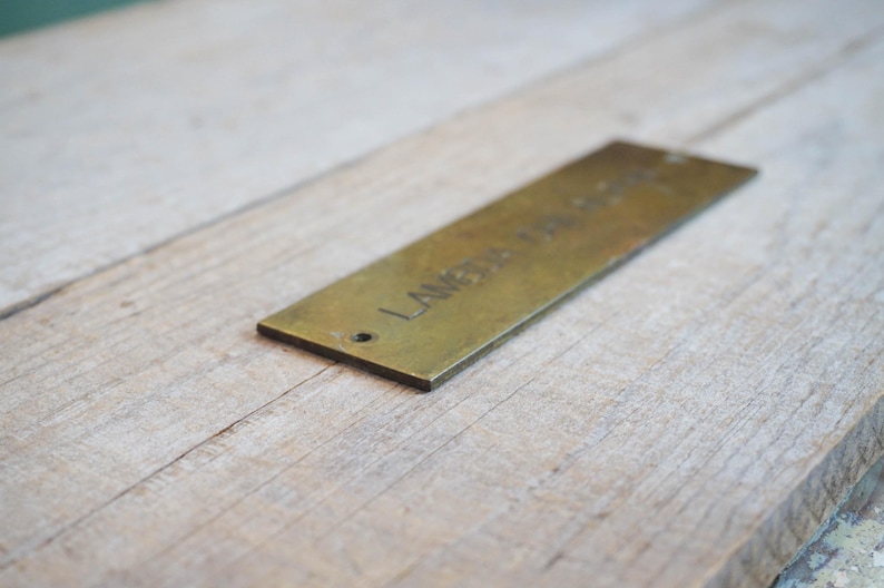 Pode incluir: Uma etiqueta retangular de lat&atilde;o com o texto gravado "LAMBDA DESIGN" e um pequeno furo numa extremidade. A etiqueta est&aacute; colocada sobre uma superf&iacute;cie de madeira clara, criando um contraste.