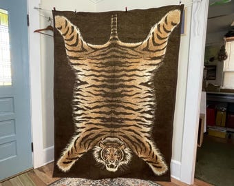 Pode incluir: Um tapete marrom retangular grande com um design de pele de tigre. O tapete apresenta uma representação detalhada do corpo de um tigre, incluindo listras, cabeça e patas. O tapete está pendurado em uma parede.