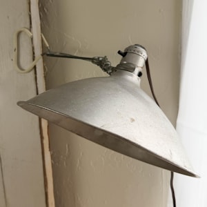 May include: A vintage, silver-toned metal lamp with a conical shade. The lamp has an adjustable arm and a brown electrical cord. The lamp is mounted on a wall and is designed to direct light downwards.