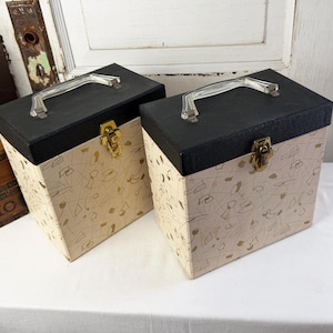May include: Two square storage cases with black lids and cream-colored bodies featuring a gold geometric line pattern. Each case has a clear plastic handle on top and a gold-toned metal latch closure on the front for securing the lid in place.
