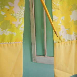 Pode incluir: Duas cortinas amarelas com um padr&atilde;o floral em tons de verde, branco e amarelo. As cortinas t&ecirc;m uma guarni&ccedil;&atilde;o de renda branca e um fundo amarelo s&oacute;lido.