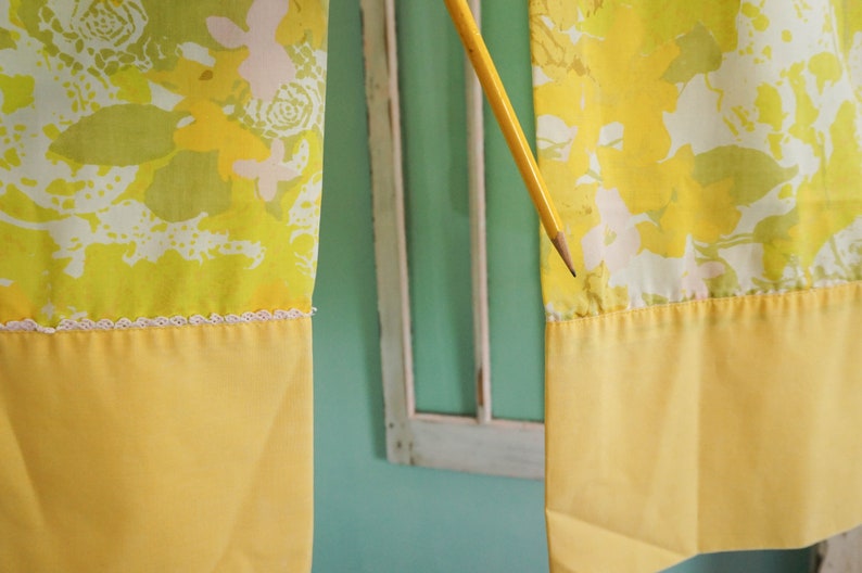 Pode incluir: Duas cortinas amarelas com um padr&atilde;o floral em tons de verde, branco e amarelo. As cortinas t&ecirc;m uma guarni&ccedil;&atilde;o de renda branca e um fundo amarelo s&oacute;lido.