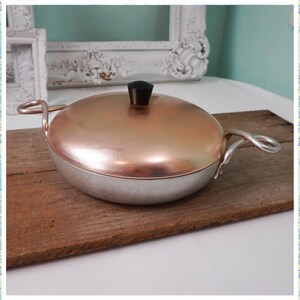 May include: A copper and aluminum casserole dish with a black knob on the lid. The dish is sitting on a wooden cutting board.