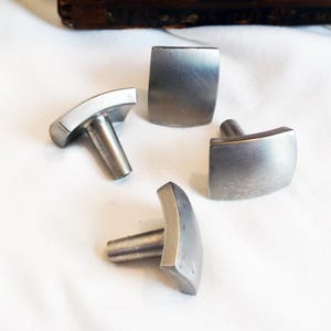 May include: Four silver-toned cufflinks with a brushed metal finish. Two cufflinks have a rectangular shape, while the other two have a curved, rectangular design. The cufflinks are arranged on a white surface.