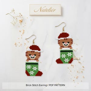 Peut inclure: Une paire de boucles d'oreilles en perles représentant un chat marron portant un bonnet de Père Noël rouge et blanc, niché dans une chaussette de Noël verte à motifs de flocons blancs. Crochets dorés. Texte Natelier visible.