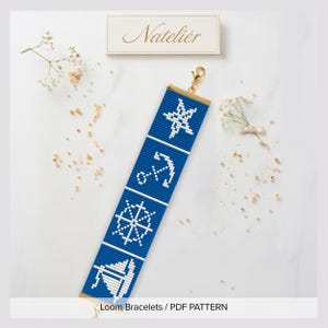 Puede incluir: Una pulsera tejida en azul y blanco con un patrón náutico de una estrella, un ancla, un timón y un velero. La pulsera tiene un cierre de metal dorado en la parte superior. Arriba hay una etiqueta rectangular con el texto Natelier.