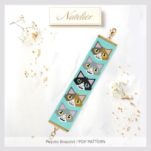 Könnte beinhalten: Ein Perlenarmband mit hellblauem Hintergrund und einem wiederkehrenden Muster aus fünf stilisierten Katzengesichtern in verschiedenen Farben wie Braun, Weiß, Grau und Schwarz. Das Armband hat goldfarbene Metallendkappen und einen Karabinerverschluss.