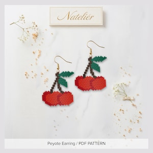 May include: A pair of beaded cherry earrings featuring red fruit, brown stems, and green leaves crafted with a peyote stitch pattern. The earrings hang from gold-toned hooks. A rectangular label at the top displays the text Nateliér.
