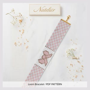 Könnte beinhalten: Ein Webarmband-Muster mit einem sich wiederholenden Gitterdesign in Weiß- und Zartrosatönen sowie einem zentralen Motiv in Form eines kleinen rosa Tieres. Das Armband ist mit einem goldfarbenen Metallverschluss und dem Schriftzug Natelier versehen.