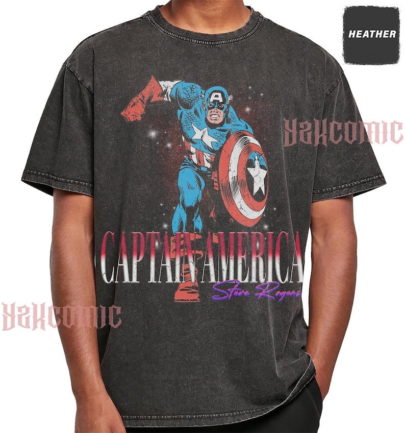 May include: A charcoal grey heather t-shirt featuring a graphic of Captain America in a dynamic pose with a shield. The text CAPTAIN AMERICA is printed in white and red, with Steve Rogers in purple script below. The word HEATHER appears in a small box.