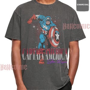 May include: A charcoal gray short-sleeve t-shirt featuring a graphic of Captain America in a dynamic pose with a shield. The text CAPTAIN AMERICA is printed in large white letters, with Steve Rogers in purple script below. The fabric has a washed finish.