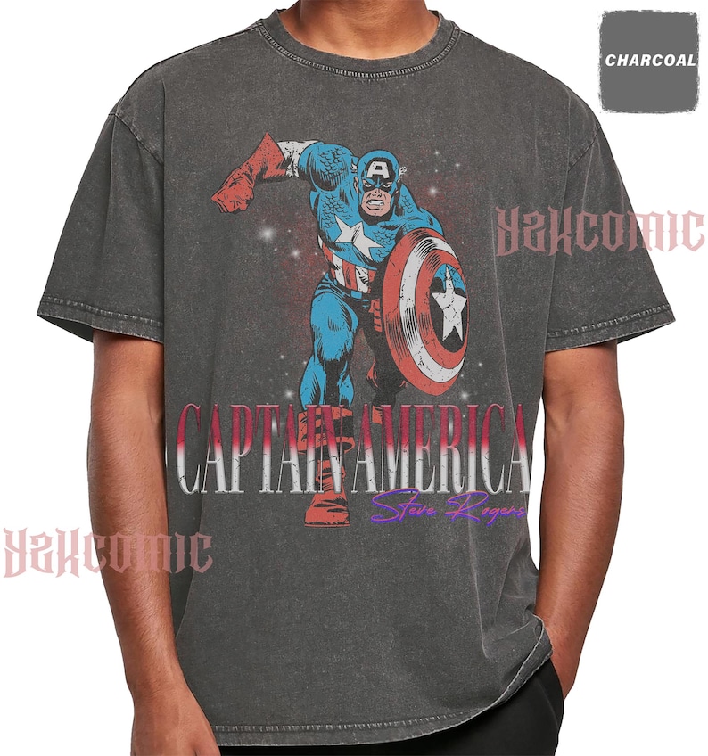May include: A charcoal gray short-sleeve t-shirt featuring a graphic of Captain America in a dynamic pose with a shield. The text CAPTAIN AMERICA is printed in large white letters, with Steve Rogers in purple script below. The fabric has a washed finish.