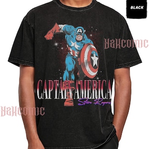 May include: A black acid-wash t-shirt featuring a graphic of Captain America in a dynamic pose with a shield. The text CAPTAIN AMERICA and Steve Rogers is printed below the character. A small black box in the corner contains the word BLACK.