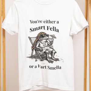 May include: White t-shirt with the text "You're either a Smart Fella or a Fart Smella." The graphic shows a frog in a chair, wearing sunglasses and a hat, holding a drink. The shirt is hanging on a wooden clothing rack.