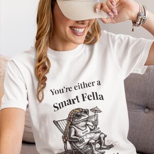 May include: White t-shirt with the text "You're either a Smart Fella or a Fart Smella" and a cartoon frog wearing sunglasses, a dress, and holding a drink. The frog is sitting in a beach chair. A beige baseball cap is worn.