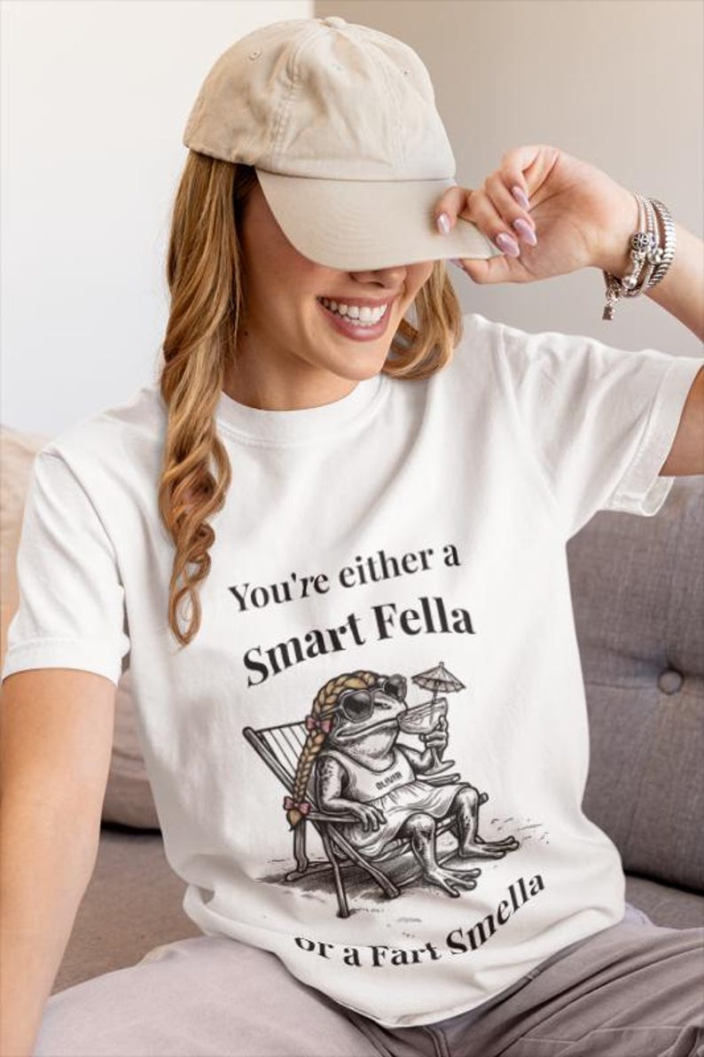 May include: White t-shirt with the text "You're either a Smart Fella or a Fart Smella" and a cartoon frog wearing sunglasses, a dress, and holding a drink. The frog is sitting in a beach chair. A beige baseball cap is worn.