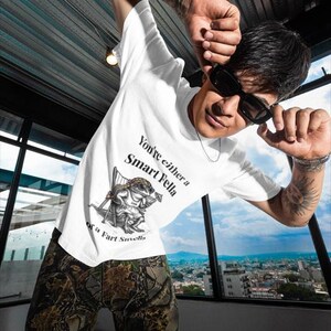 May include: White t-shirt with the text "You're either a Smart Fella or a Fart Smella" and a cartoon frog illustration. The person is wearing camouflage pants and sunglasses. The background shows a cityscape.