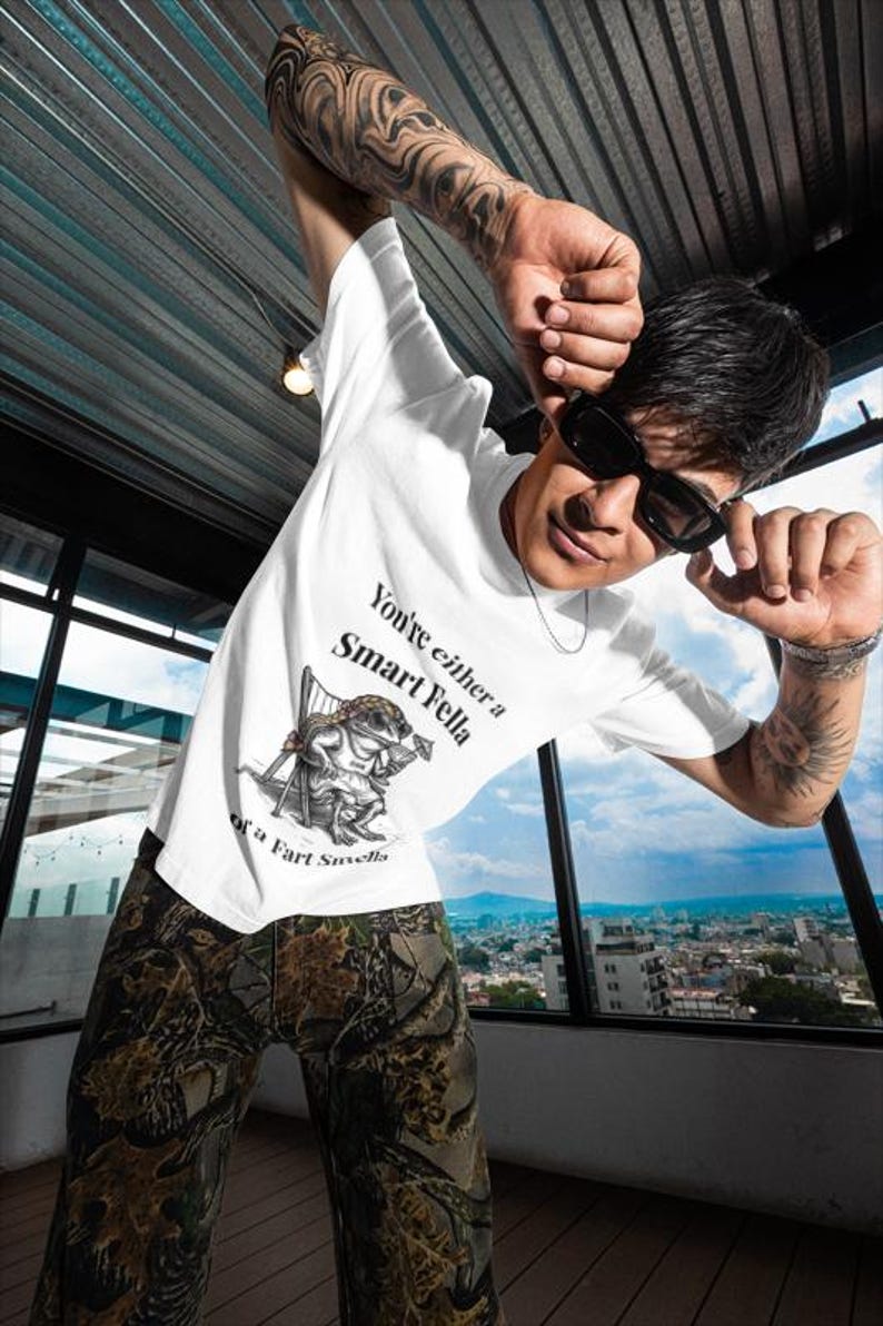 May include: White t-shirt with the text "You're either a Smart Fella or a Fart Smella" and a cartoon frog illustration. The person is wearing camouflage pants and sunglasses. The background shows a cityscape.