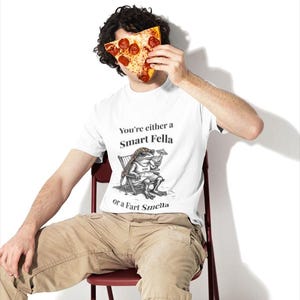 May include: White t-shirt with the text "You're either a Smart Fella or a Fart Smella" and a cartoon frog illustration. The person is holding a slice of pizza with pepperoni. The person is wearing khaki pants and sitting on a red chair.