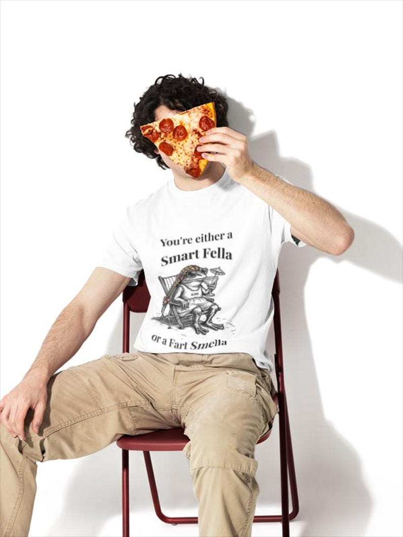 May include: White t-shirt with the text "You're either a Smart Fella or a Fart Smella" and a cartoon frog illustration. The person is holding a slice of pizza with pepperoni. The person is wearing khaki pants and sitting on a red chair.