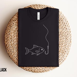 May include: A black t-shirt featuring a simple white line art graphic of a fish swimming towards a fishing hook and line. The shirt has a crew neckline and a white brand label visible on the inside collar. The design is centred on the chest.