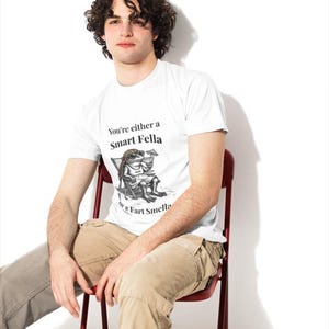 May include: White t-shirt with the text "You're either a Smart Fella or a Fart Smella" and a cartoon illustration. The person is wearing khaki pants and sitting on a red chair. The background is white.