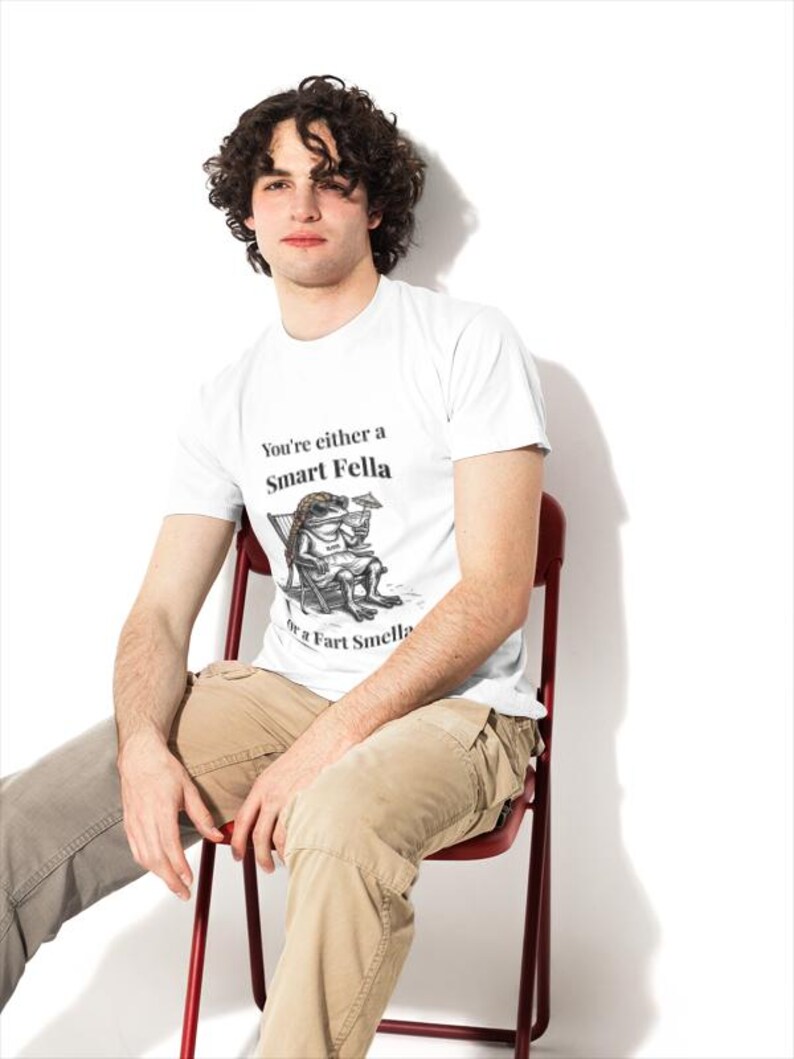 May include: White t-shirt with the text "You're either a Smart Fella or a Fart Smella" and a cartoon illustration. The person is wearing khaki pants and sitting on a red chair. The background is white.