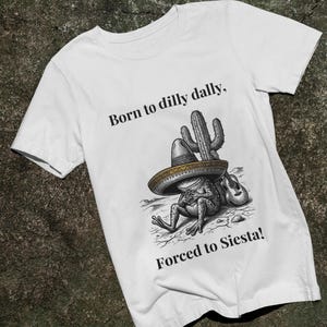 Puede incluir: Camiseta blanca con un dise&ntilde;o de una rana con sombrero apoyada en un cactus. El texto dice "Born to dilly dally, Forced to Siesta!" en negro. El dise&ntilde;o incluye una guitarra.