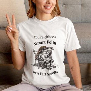 May include: White t-shirt with the text "You're either a Smart Fella or a Fart Smella." The shirt features a cartoon frog wearing sunglasses and shorts, relaxing in a beach chair and holding a cocktail. The person is sitting on a gray couch.