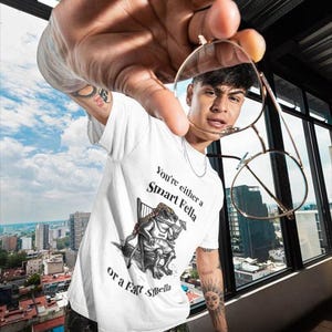 May include: White t-shirt with the text "You're either a Smart Fella or a Fart Smella" and a cartoon frog illustration. The person is holding gold-rimmed glasses. The background shows a cityscape.
