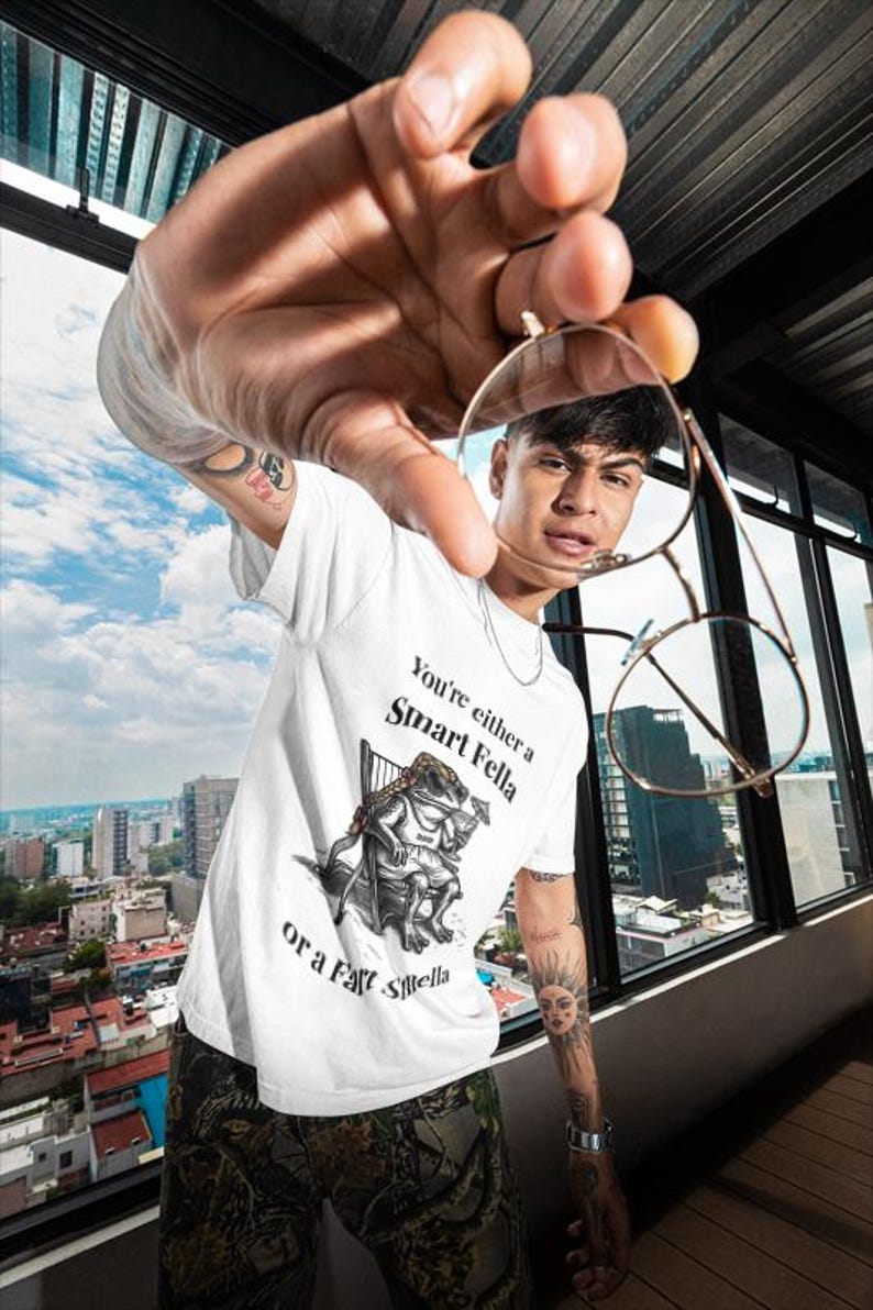 May include: White t-shirt with the text "You're either a Smart Fella or a Fart Smella" and a cartoon frog illustration. The person is holding gold-rimmed glasses. The background shows a cityscape.