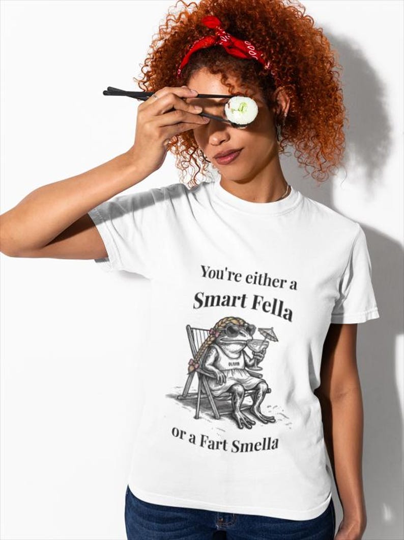 May include: White t-shirt with the text "You're either a Smart Fella or a Fart Smella" and a cartoon frog wearing sunglasses, sitting in a beach chair, holding a drink. The person is holding sushi with chopsticks.