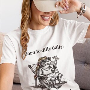 May include: White t-shirt with a graphic of a frog in sunglasses, sitting in a beach chair and holding a cocktail. The text "Born to dilly dally, Forced to Beach!" is printed on the shirt. A beige baseball cap is also visible.