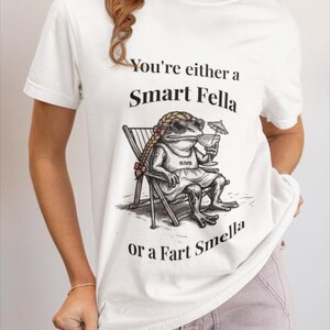 May include: White t-shirt with the text "You're either a Smart Fella or a Fart Smella." The shirt features a cartoon frog wearing sunglasses and a dress, sitting in a beach chair and holding a cocktail.