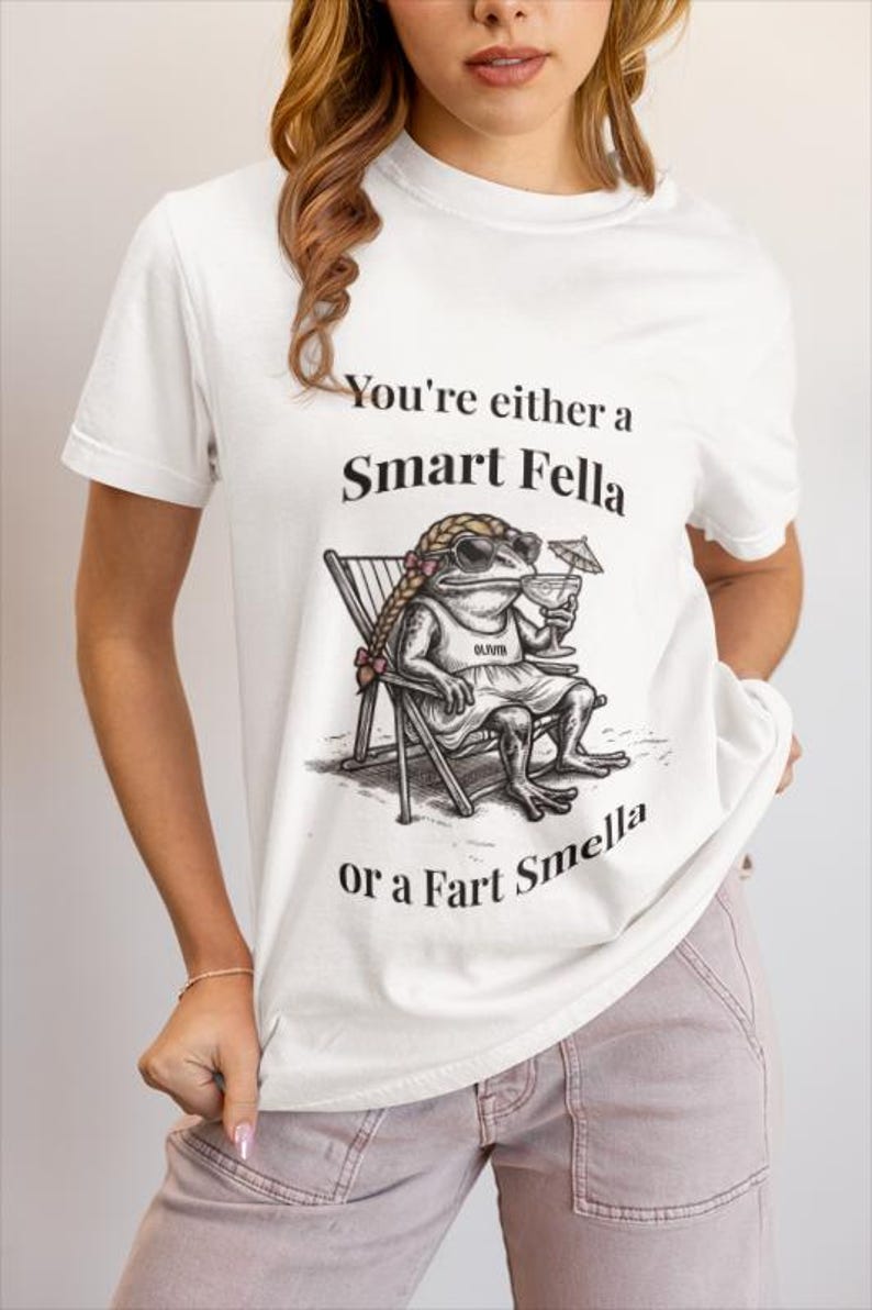 May include: White t-shirt with the text "You're either a Smart Fella or a Fart Smella." The shirt features a cartoon frog wearing sunglasses and a dress, sitting in a beach chair and holding a cocktail.