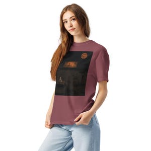 May include: A dusty rose short-sleeved t-shirt featuring a large rectangular graphic print on the chest. The graphic depicts a dark, moody scene with a small figure standing at a table, a glowing orange moon, and a distant illuminated structure.