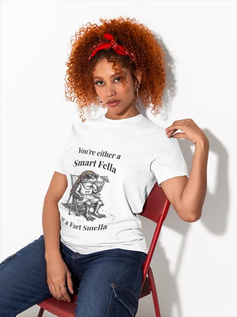 May include: White t-shirt with the text "You're either a Smart Fella or a Fart Smella" and a cartoon frog illustration. The frog is sitting in a chair, wearing shorts and holding a drink. The person is wearing blue jeans and a red headband.