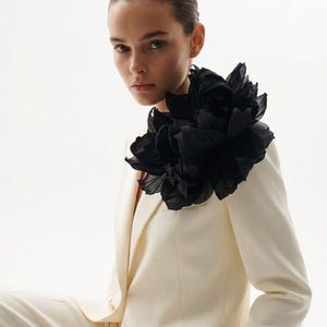 May include: A cream-coloured suit with a large black floral accent. The suit jacket has a tailored fit, and the black floral piece is positioned on the shoulder. The background is plain white.