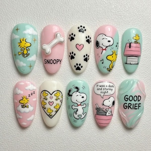May include: A set of ten artificial nails with cartoon character designs. The nails feature Snoopy, Woodstock, paw prints, hearts, and text. Colors include pink, white, blue, and yellow. The nails are oval-shaped and have a glossy finish.