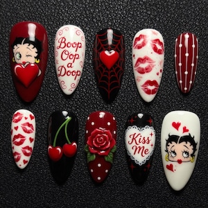 May include: A collection of ten artificial nails with various designs. The designs include Betty Boop, lips, hearts, cherries, a rose, and the text "Boop Oop a Doop" and "Kiss Me". The nails are in shades of red, black, and white.