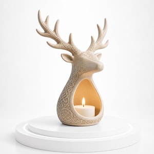 May include: A beige deer-shaped candle holder with detailed Celtic knotwork and floral designs. The deer features large antlers and a cavity for a lit candle. The candle holder is displayed on a white, tiered platform.