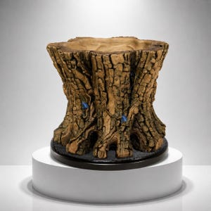 Puede incluir: Una maceta marrón con forma de tocón de árbol, con un diseño de corteza texturizada. La maceta tiene una base oscura y detalles azules. Se encuentra sobre un pedestal blanco, ideal para decoración interior o exterior.