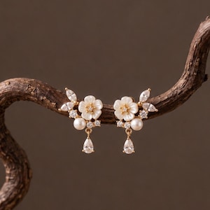 Può includere: Un paio di orecchini color oro con motivi floreali bianchi, piccole perle rotonde e cristalli trasparenti a forma di marquise e goccia. Gli orecchini presentano un design a grappolo con un cristallo pendente che termina a goccia.