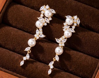 Puede incluir: Un par de pendientes colgantes en tono dorado con una disposición vertical de perlas blancas redondas y piedras de circonita cúbica transparentes en talla marquesa. El diseño alterna perlas y piedras brillantes en una estructura fina y alargada.